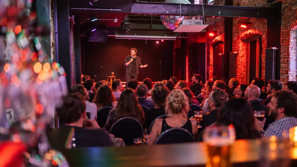 Blick über die Theke auf die Bühne, das Publikum lacht mit Bier in der Hand