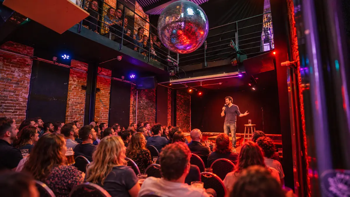 Comedian auf der Bühne unter der Discokugel, Gäste auf der Galerie und in den Stuhlreihen lachen
